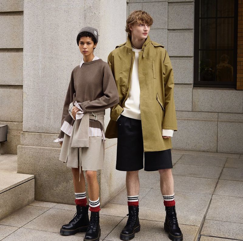 Two people in outerwear and boots stand against a building. One wears a coat and the other a sweater with skirt and beanie.