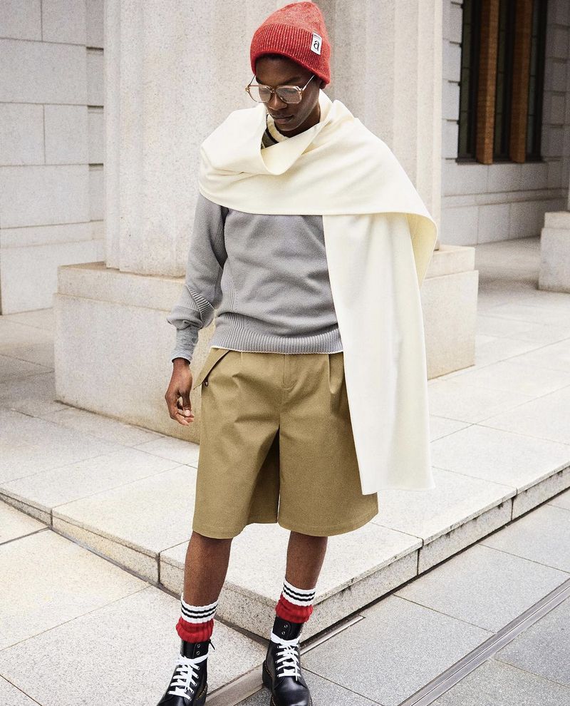 Model Odinaka Ekezie in a red beanie, cream draped wrap, grey sweater, and khaki shorts standing outdoors near classical stone columns.