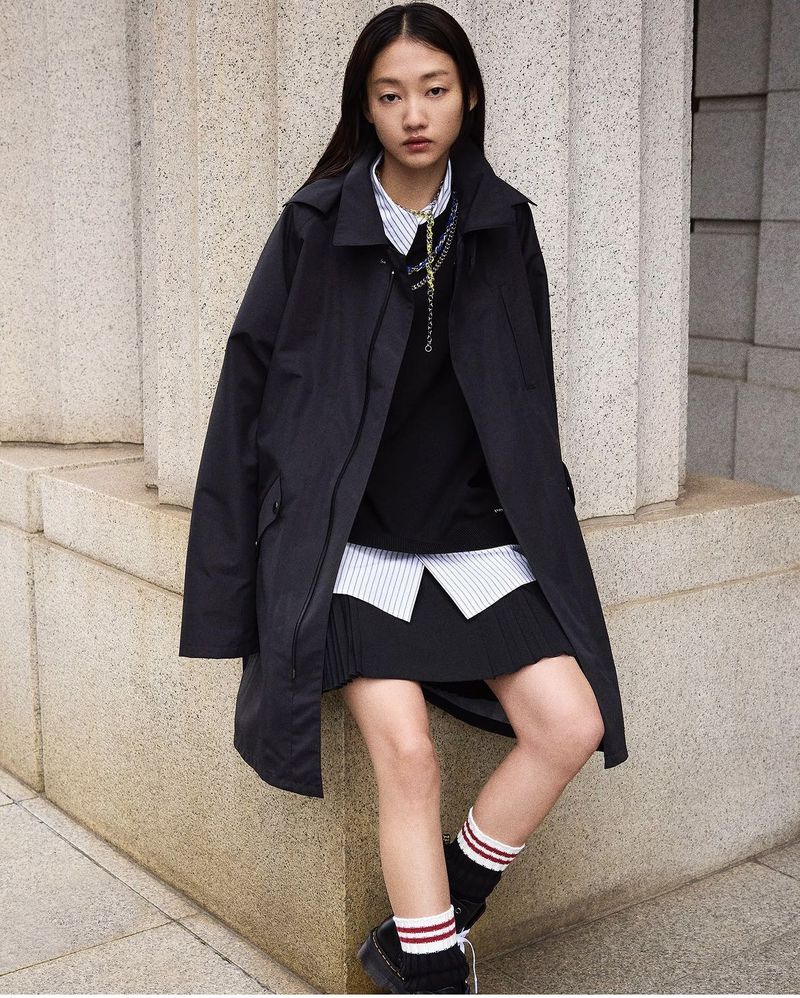 An Asian woman poses outdoors with a black coat, layered top, skirt, socks, and platform shoes against a concrete wall in a medium shot.