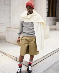 Model Odinaka Ekezie in a red beanie, cream draped wrap, grey sweater, and khaki shorts standing outdoors near classical stone columns.
