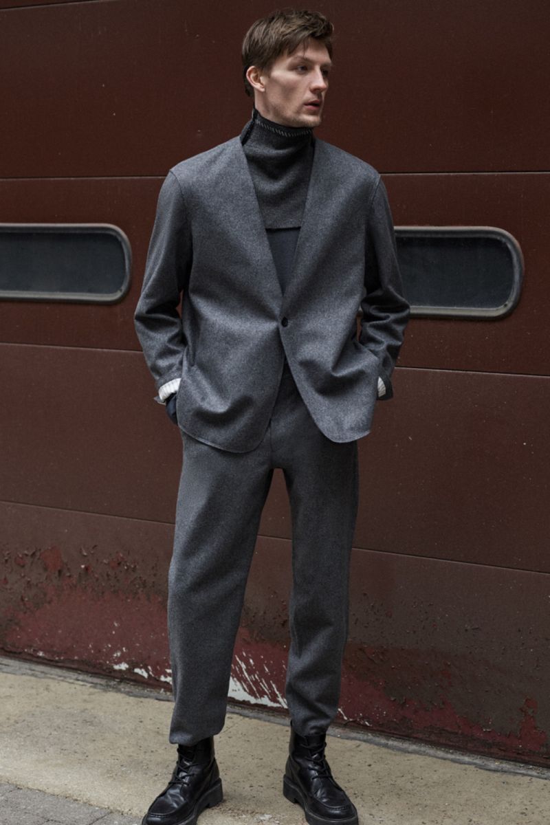 A stylish adult male model poses in a gray suit and black boots against a brown wall in an urban setting for a fashion editorial shot.