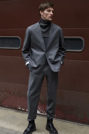A stylish adult male model poses in a gray suit and black boots against a brown wall in an urban setting for a fashion editorial shot.