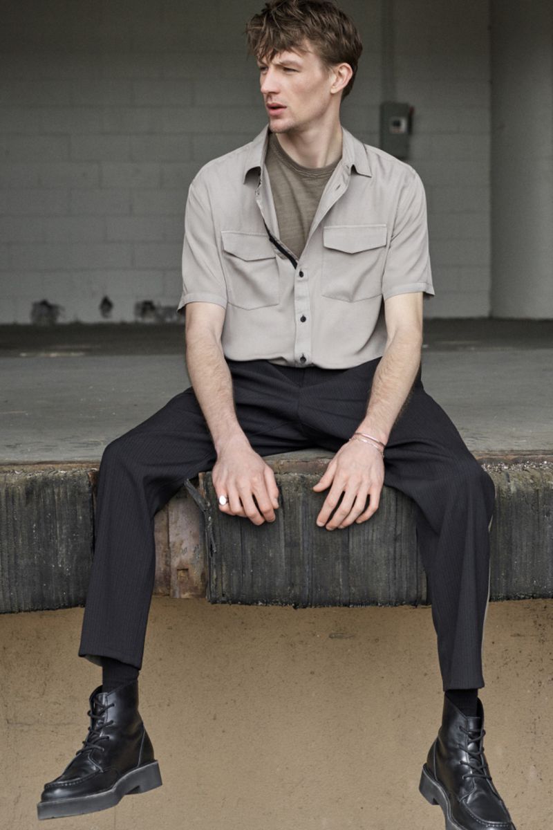 Young adult male in short-sleeved shirt and dark pants sits with legs dangling near a concrete wall, wearing black boots.