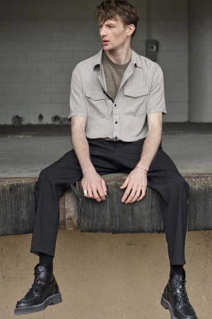 Young adult male in short-sleeved shirt and dark pants sits with legs dangling near a concrete wall, wearing black boots.