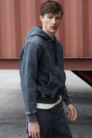 Adult man in denim hoodie and jeans posing outdoors against red metal wall in medium studio shot, looking at the camera with neutral expression.