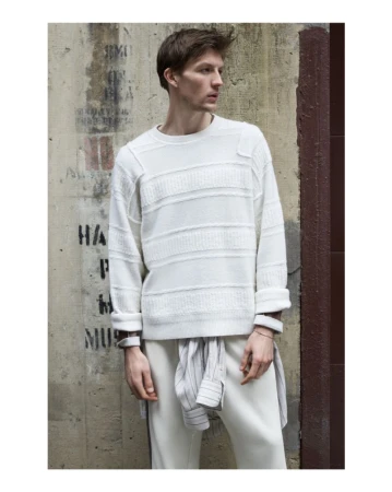 Man in white textured sweater and pants posing against a concrete wall with stenciled writing and a dark brown corner accent.