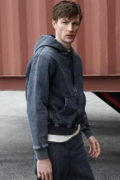 Model Finnlay Davis wearing a washed denim hoodie and matching jeans against a rust red industrial shipping container.