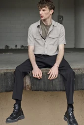 Model Finnlay Davis wearing a light grey short-sleeve shirt, black trousers, and black combat boots seated on a concrete loading dock.