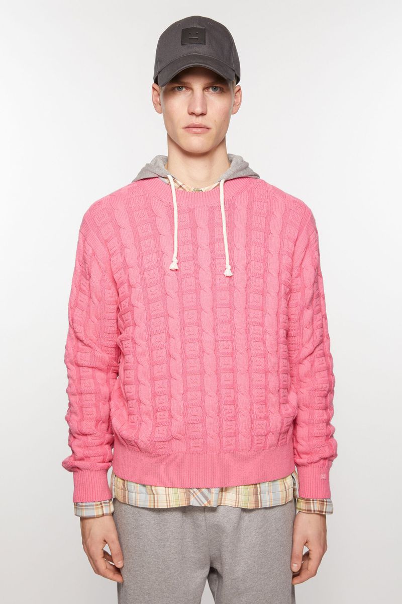 Studio shot of a young adult male wearing a pink textured hooded sweatshirt and a dark baseball cap against a white backdrop. 