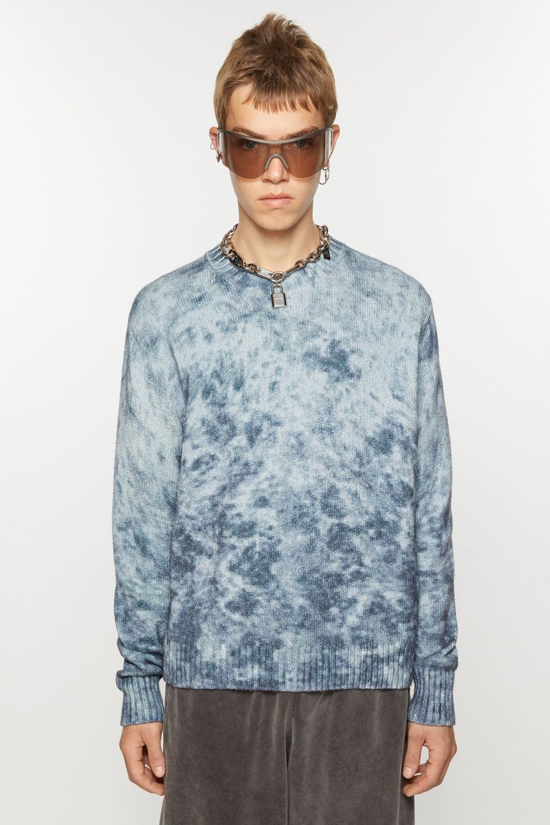 Man in a blue-and-white cloud-pattern sweater, gray shorts, sunglasses, and a silver chain posing in front of a white backdrop.