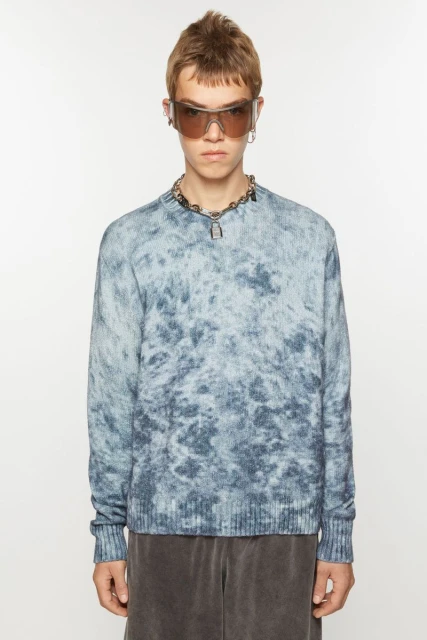Man in a blue-and-white cloud-pattern sweater, gray shorts, sunglasses, and a silver chain posing in front of a white backdrop.