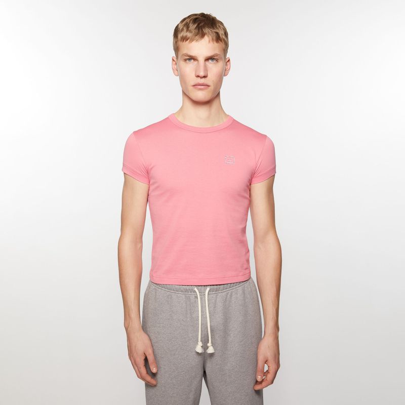 A light-skinned adult male poses indoors wearing a pink short-sleeve shirt and grey sweatpants against a white backdrop in a studio shot.