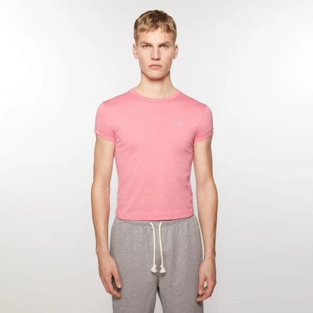 A light-skinned adult male poses indoors wearing a pink short-sleeve shirt and grey sweatpants against a white backdrop in a studio shot.