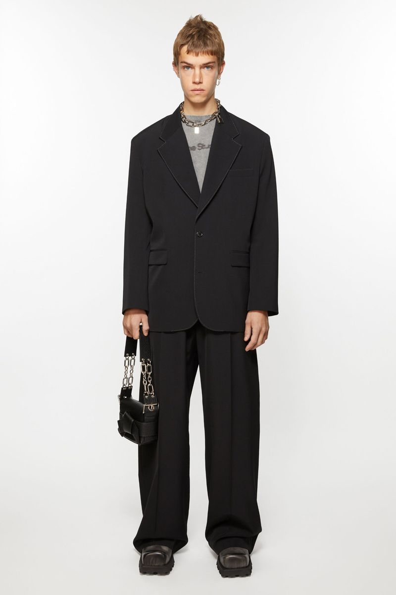 Full-length studio shot of a male model wearing a black blazer, t-shirt, black trousers, chunky shoes and carrying a black purse with chain.