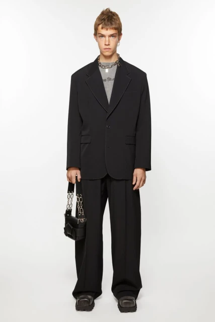 Full-length studio shot of a male model wearing a black blazer, t-shirt, black trousers, chunky shoes and carrying a black purse with chain.