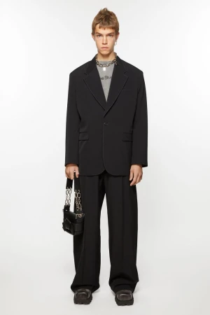 Full-length studio shot of a male model wearing a black blazer, t-shirt, black trousers, chunky shoes and carrying a black purse with chain.