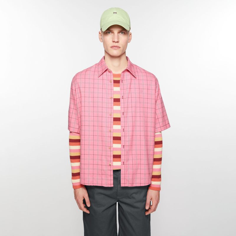 Man in pink plaid shirt and grey pants with light green cap stands against a white backdrop in a fashion studio shot.