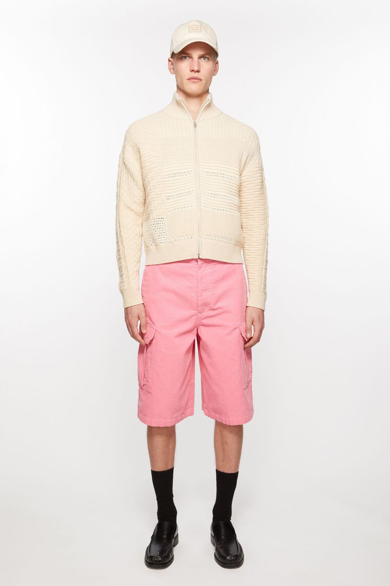 A fair-skinned young adult male poses in a studio wearing a cream-colored knit jacket, pink shorts, a matching baseball cap, black socks, and black dress shoes.
