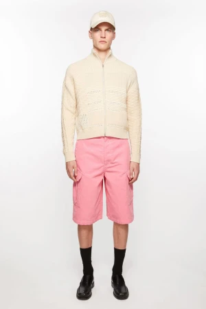 A fair-skinned young adult male poses in a studio wearing a cream-colored knit jacket, pink shorts, a matching baseball cap, black socks, and black dress shoes.
