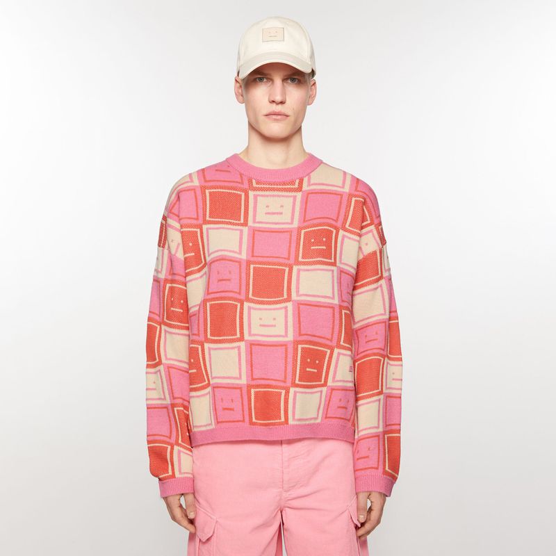 Adult male fashion model posing in a studio wearing a pink patterned sweater, pink shorts, and a beige baseball cap against a white backdrop.