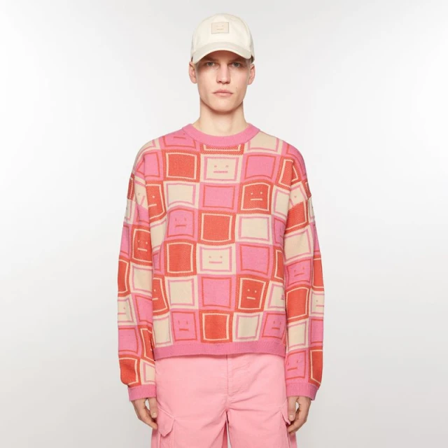 Adult male fashion model posing in a studio wearing a pink patterned sweater, pink shorts, and a beige baseball cap against a white backdrop.