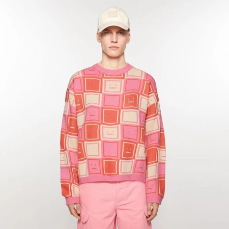 Adult male fashion model posing in a studio wearing a pink patterned sweater, pink shorts, and a beige baseball cap against a white backdrop.