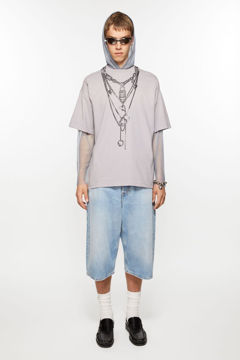 Model in an oversized chain-print t-shirt, light blue denim jorts, and black loafers against a seamless white studio background.