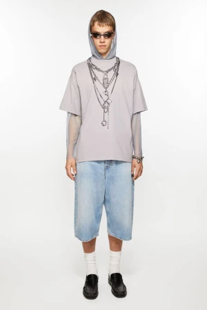 Model in an oversized chain-print t-shirt, light blue denim jorts, and black loafers against a seamless white studio background.