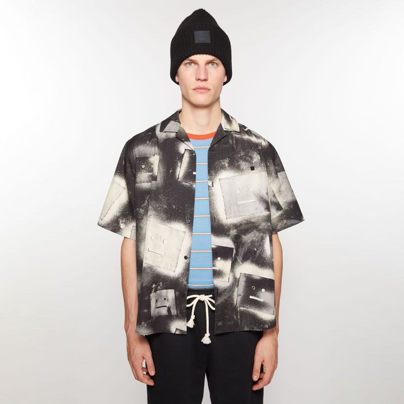 Male fashion model poses in a black and white patterned shirt, black pants, and a black beanie against a neutral background.