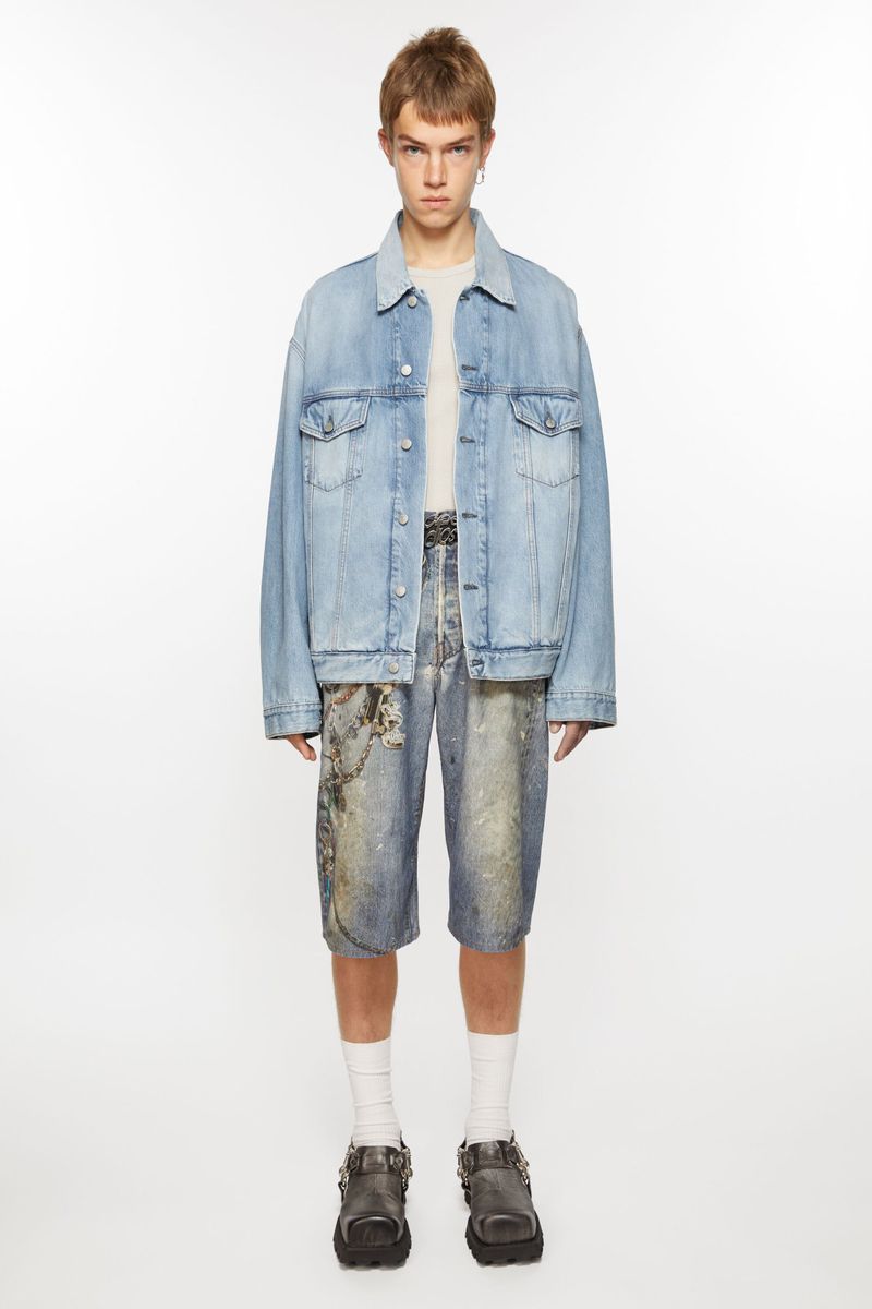 Full-length studio shot of a young adult wearing a light-wash denim jacket, printed denim shorts, white socks, and buckle-strap shoes.