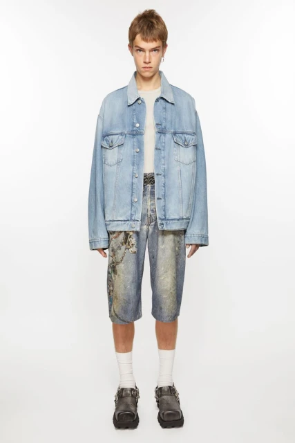 Full-length studio shot of a young adult wearing a light-wash denim jacket, printed denim shorts, white socks, and buckle-strap shoes.