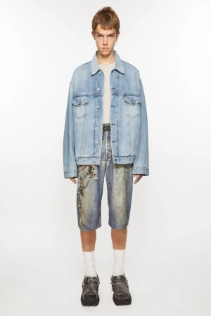 Full-length studio shot of a young adult wearing a light-wash denim jacket, printed denim shorts, white socks, and buckle-strap shoes.