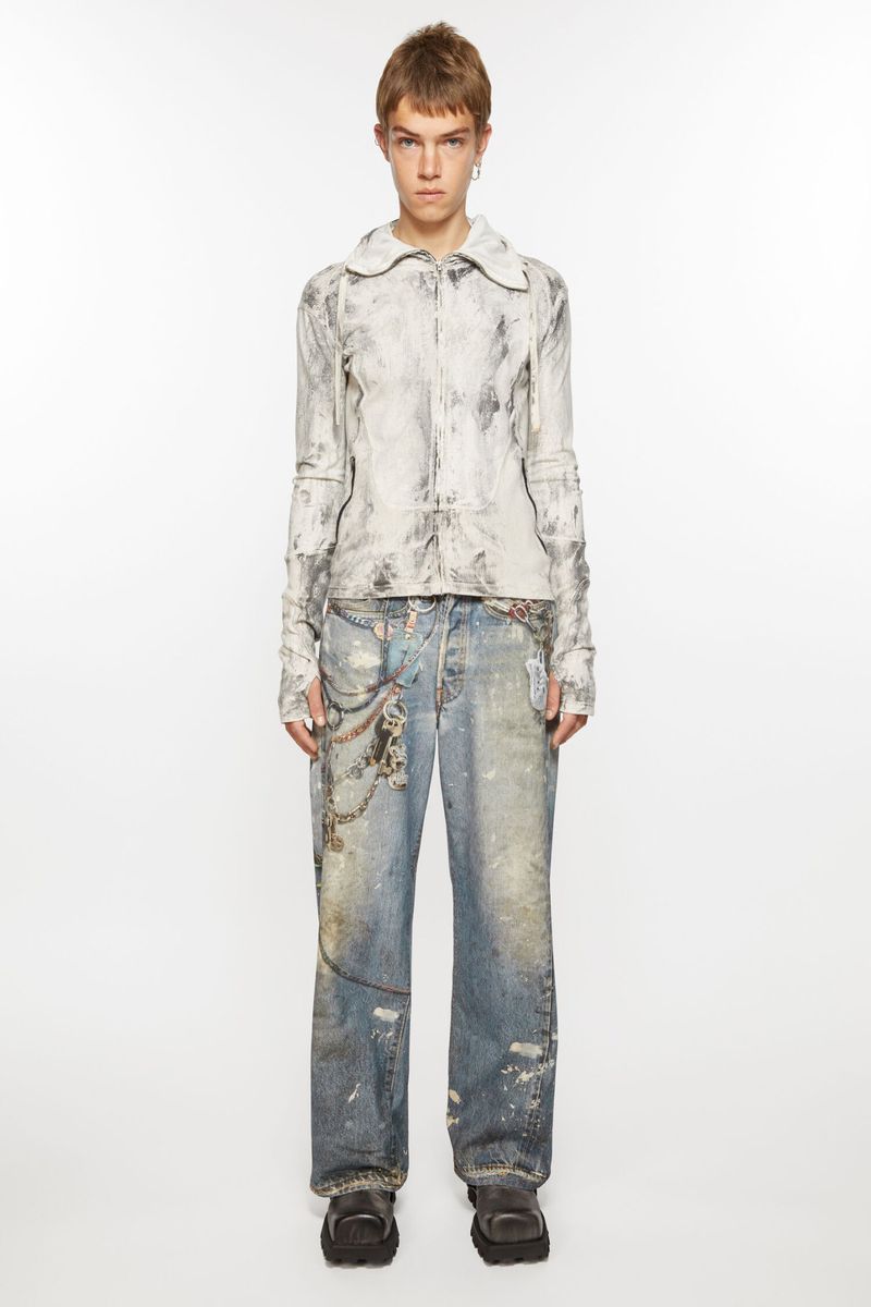 Adult female model in a full shot wearing a white marble-patterned jacket, distressed denim jeans with chains, and platform shoes on a white background.