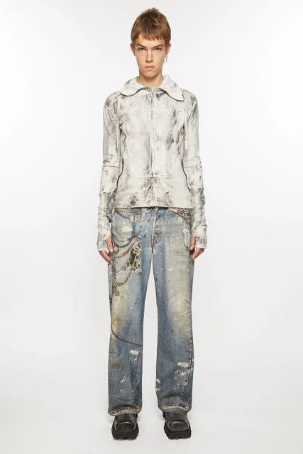 Adult female model in a full shot wearing a white marble-patterned jacket, distressed denim jeans with chains, and platform shoes on a white background.