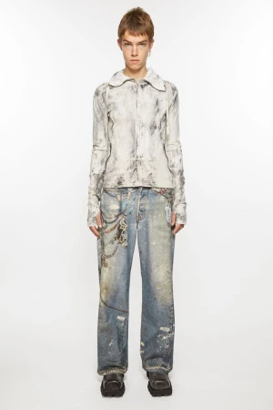 Adult female model in a full shot wearing a white marble-patterned jacket, distressed denim jeans with chains, and platform shoes on a white background.
