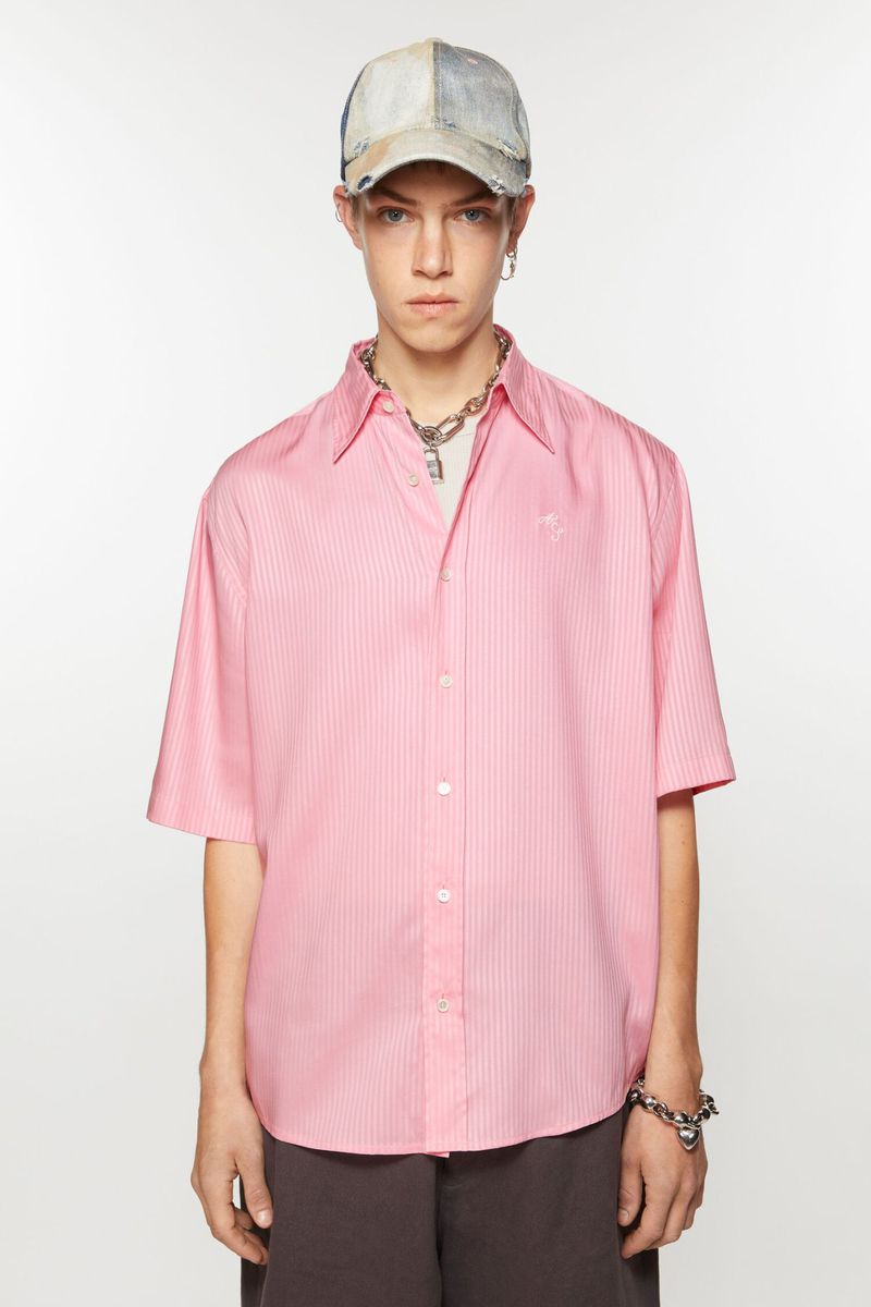 Model wearing a short-sleeve pink button-up shirt, distressed baseball cap, and silver necklace against a white seamless backdrop.