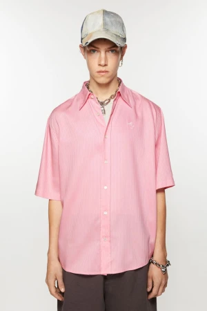 Model wearing a short-sleeve pink button-up shirt, distressed baseball cap, and silver necklace against a white seamless backdrop.