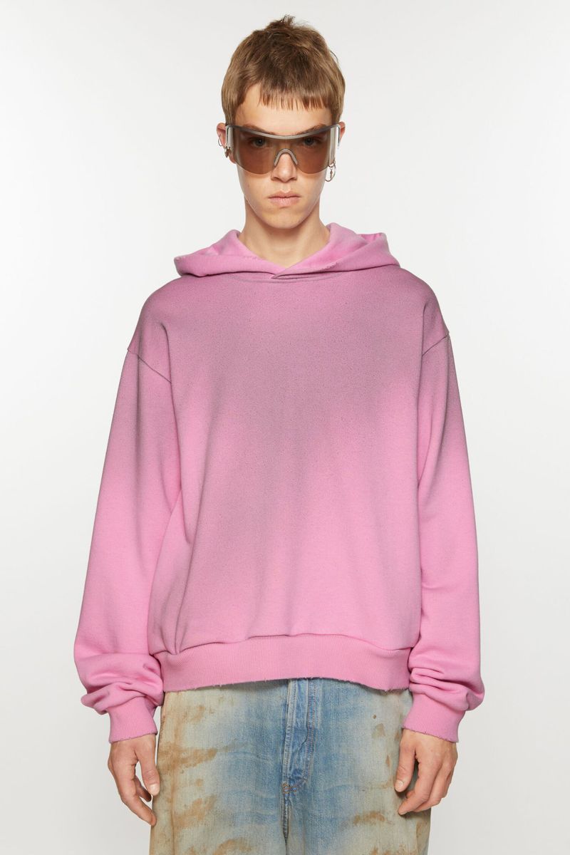 Adult male wearing a pink hoodie and sunglasses stands in a brightly lit photo studio, looking at the camera in this full shot.