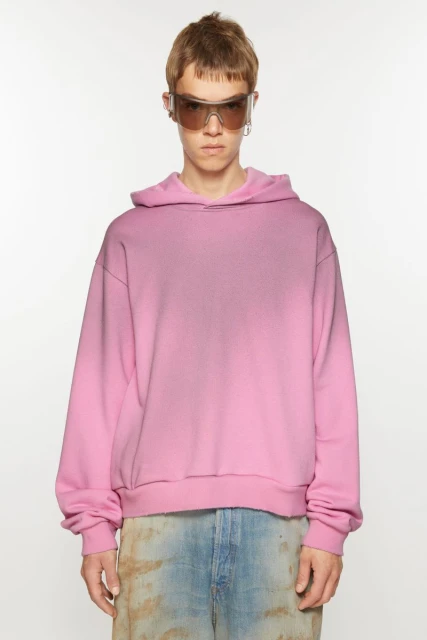 Adult male wearing a pink hoodie and sunglasses stands in a brightly lit photo studio, looking at the camera in this full shot.