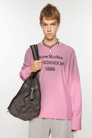 A young adult in a pink Acne Studios long-sleeve shirt with a silver necklace, grey pants, and a grey tote bag stands in a white studio.