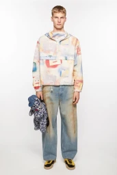 Male model standing against a white seamless background wearing a printed windbreaker, distressed wide-leg jeans, and sneakers.