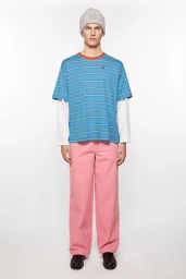 Model standing against a white seamless backdrop wearing a blue striped t-shirt, pink trousers, and a grey beanie.