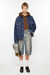 Male model wearing a dark denim jacket, brown hoodie, and paint-splattered denim shorts against a white seamless background.