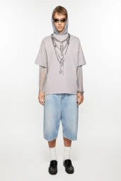 Model in an oversized chain-print t-shirt, light blue denim jorts, and black loafers against a seamless white studio background.