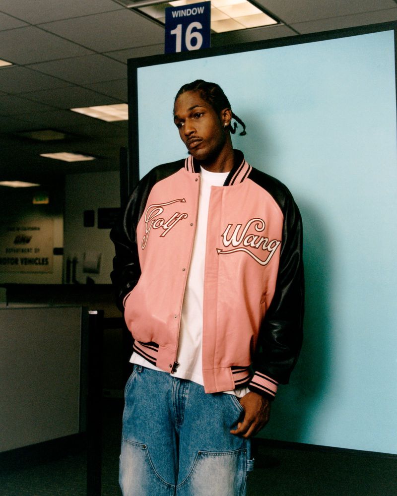 Adult male in a pink 'Golf Wang' varsity jacket and denim jeans posing in an indoor Department of Motor Vehicles environment.
