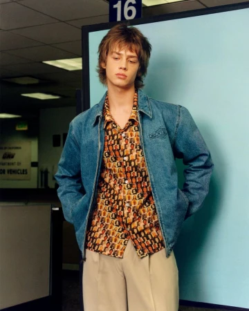 Stylish young man in printed shirt and jean jacket stands in a DMV-like environment, posing for a fashion or editorial style photograph.