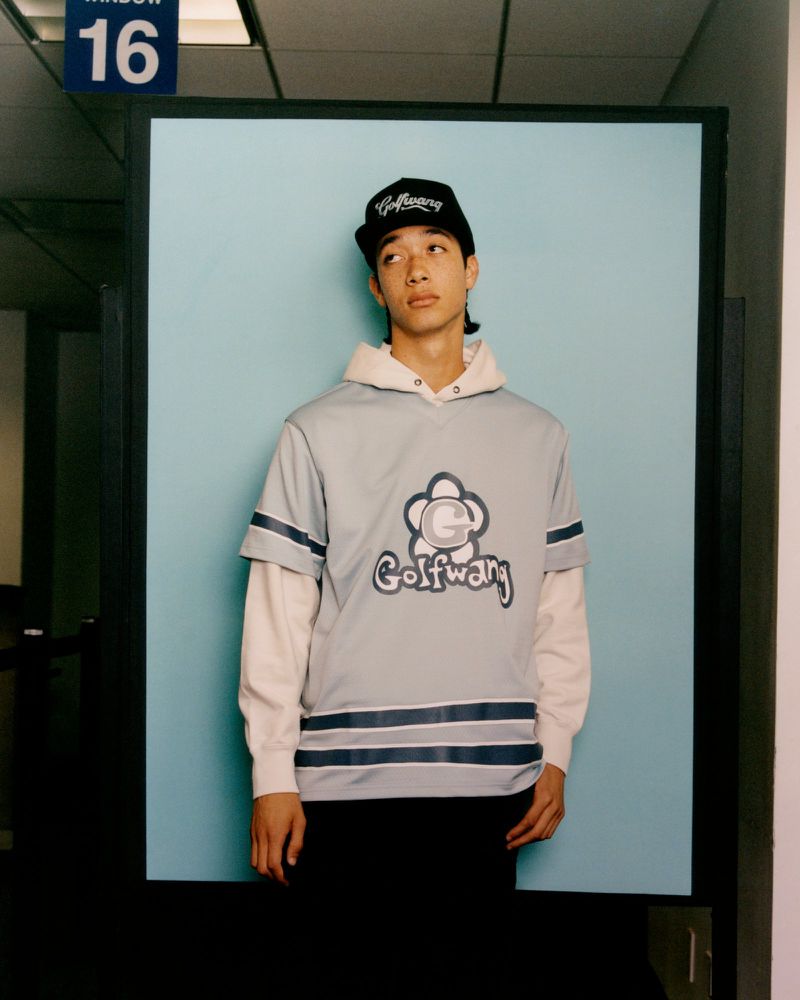 A young adult in a Golf Wang shirt and hat poses against a light blue background, creating a fashion-focused, eye-level portrait.