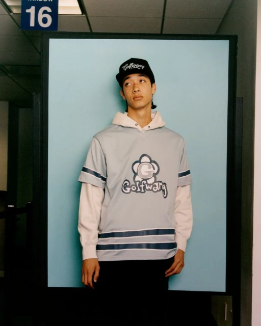 A young adult in a Golf Wang shirt and hat poses against a light blue background, creating a fashion-focused, eye-level portrait.
