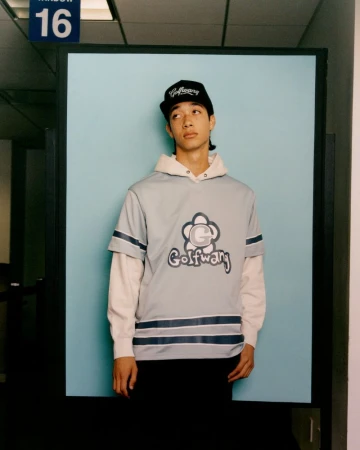 A young adult in a Golf Wang shirt and hat poses against a light blue background, creating a fashion-focused, eye-level portrait.