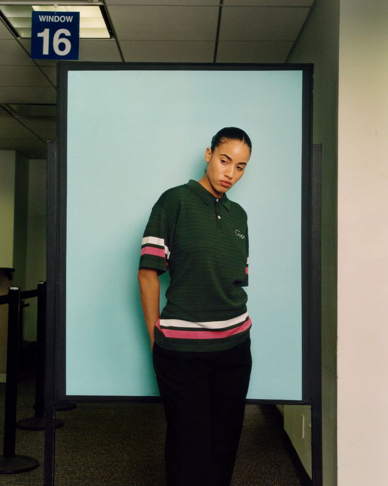 An adult female stands in a casual pose in an office setting with a blank blue sign and dark green polo shirt with white and pink stripes.